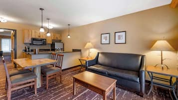 A modern living room with a wooden dining table, a black leather sofa, and a coffee table. There is a kitchen area with wooden cabinets and a microwave. Two lamps provide lighting, and the walls are decorated with framed pictures.