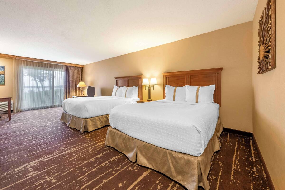 A hotel room with two beds, a large window with curtains, a wooden headboard, and a wall-mounted artwork.