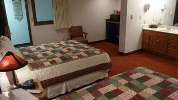A hotel room with a large bed, a smaller bed, a chair, a desk with a lamp, a mirror, and a bathroom with a sink and cabinets.