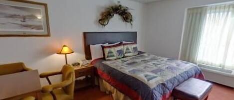 A hotel room with a bed, a desk with a lamp, a chair, and a window with curtains.
