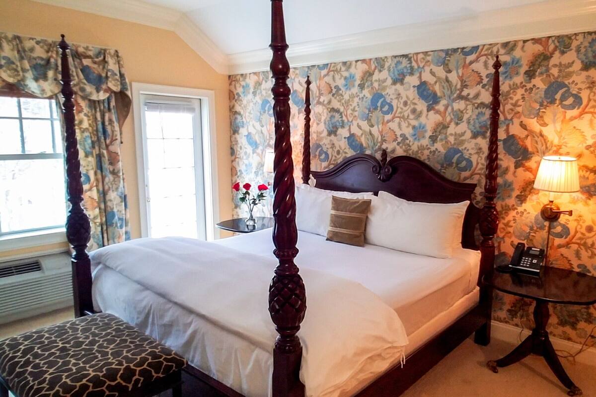 A four-poster bed with a wooden headboard, a bedside table with a lamp, and a floral wallpaper pattern.
