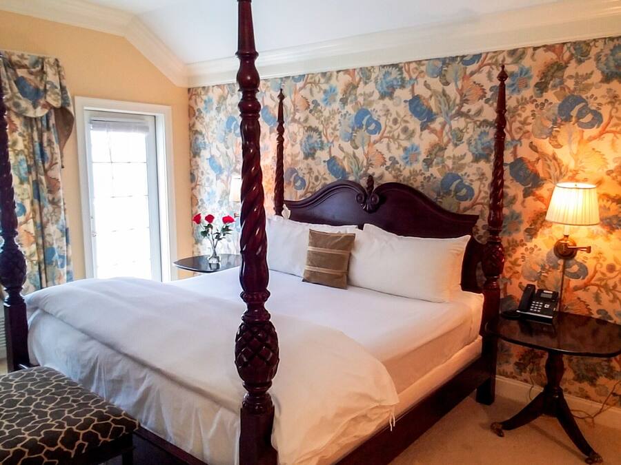A four-poster bed with a wooden headboard, a bedside table with a lamp, and a floral wallpaper pattern.