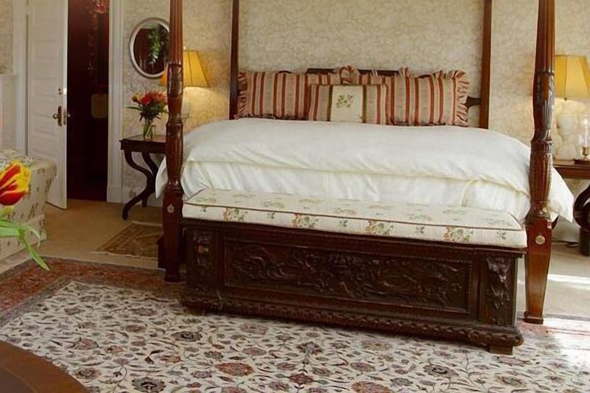 A bedroom with a four-poster bed, a fireplace, a bookshelf, and a dining table with wine glasses and a vase of flowers.