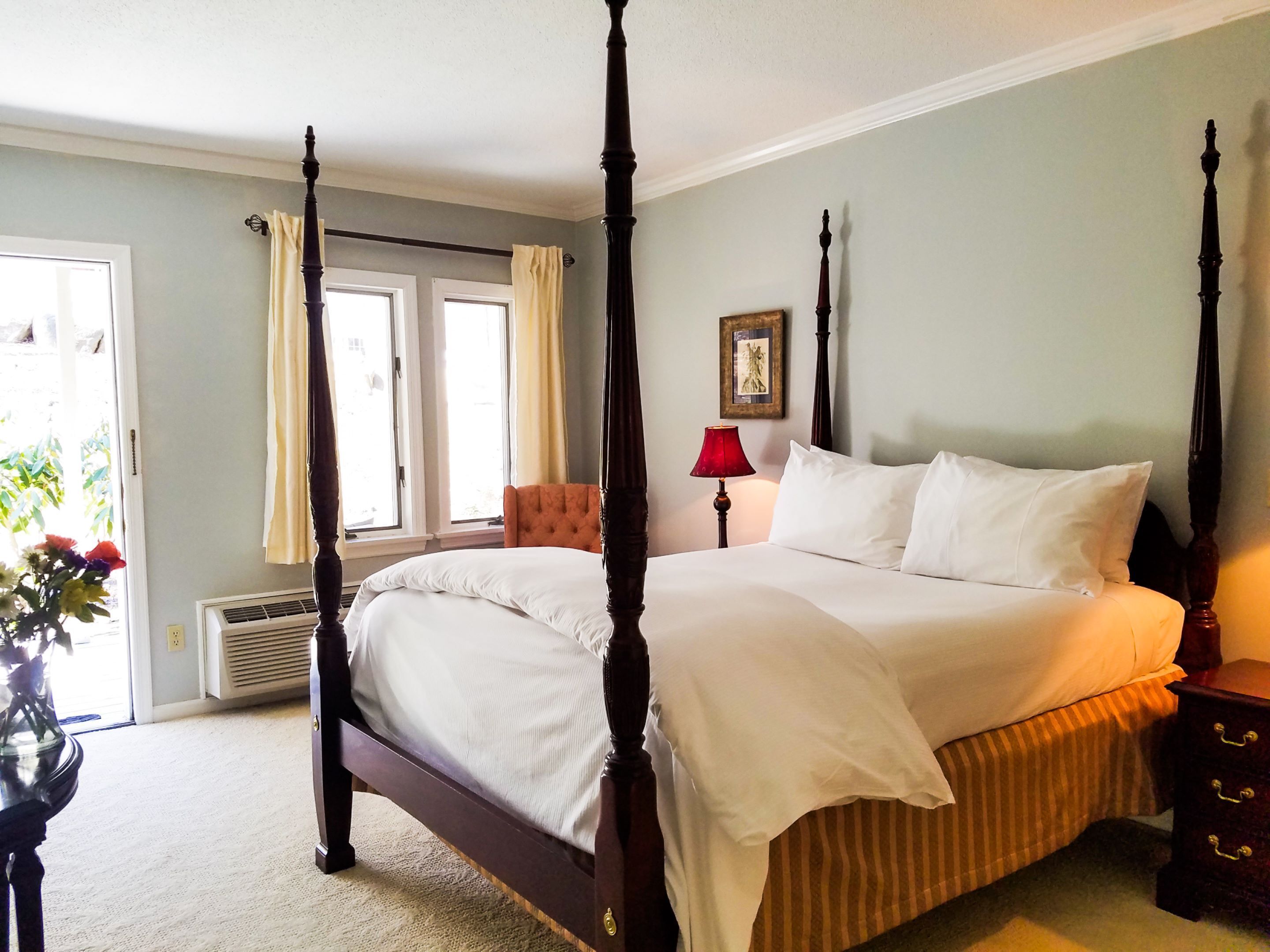 A four-poster bed with white linens, a red lamp, and a framed picture on the wall.