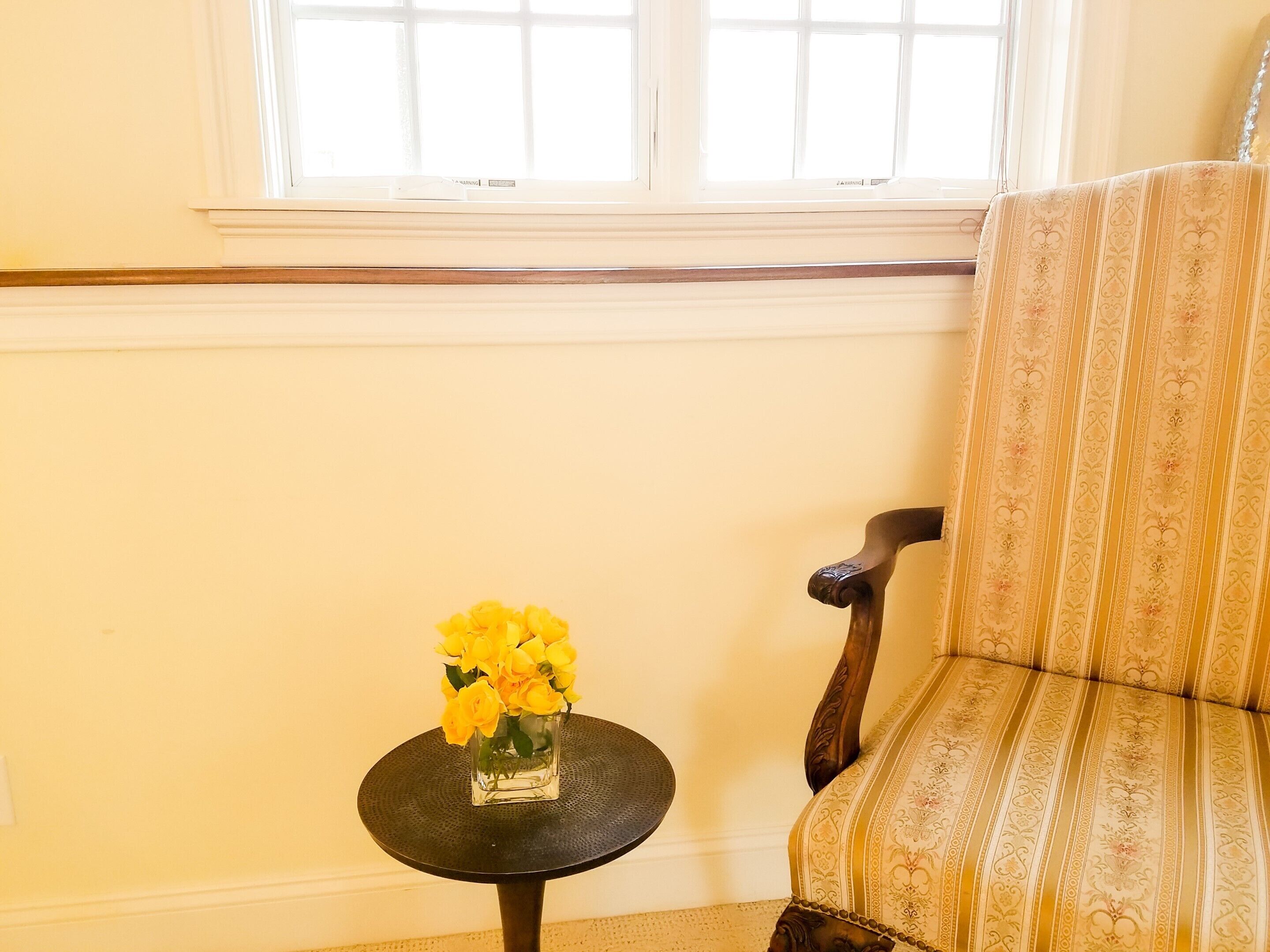 A room with a window, a chair, and a small table with a vase of yellow flowers.