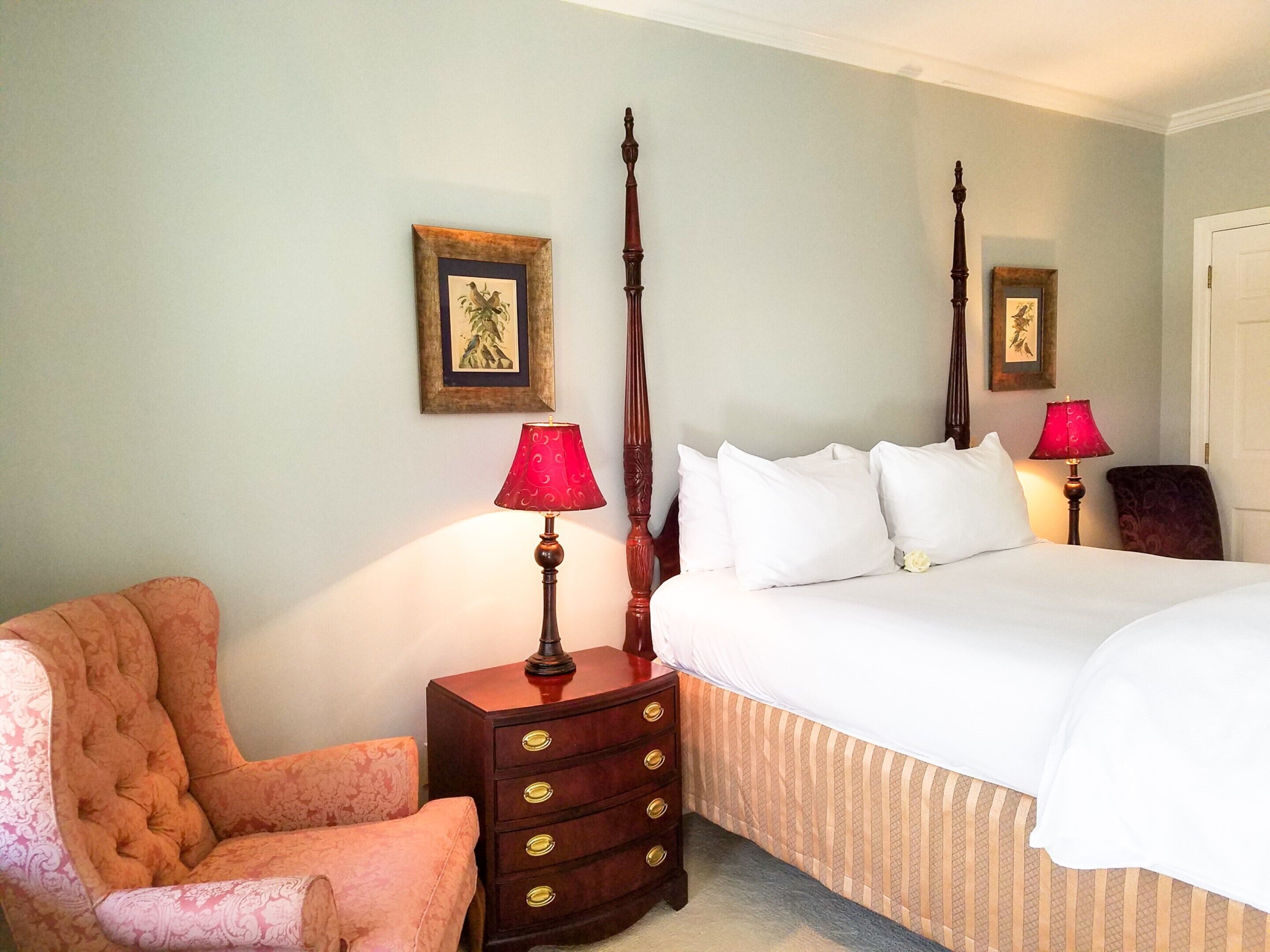 A bedroom with a four-poster bed, a wooden nightstand with a lamp, a chair, and a dresser.