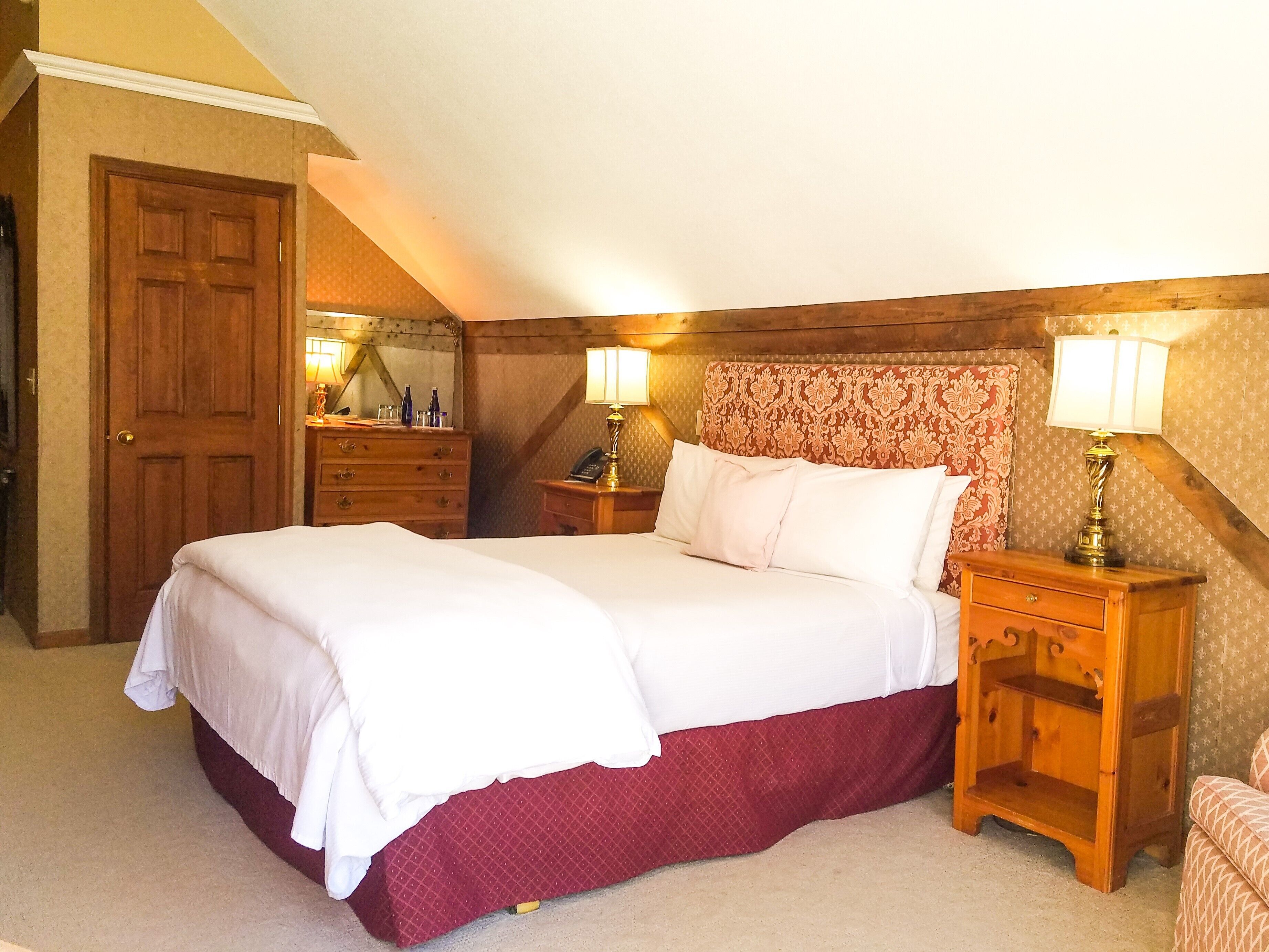 A neatly made bed with a red skirt, two bedside tables with lamps, a wooden wardrobe, and a mirror on the wall.