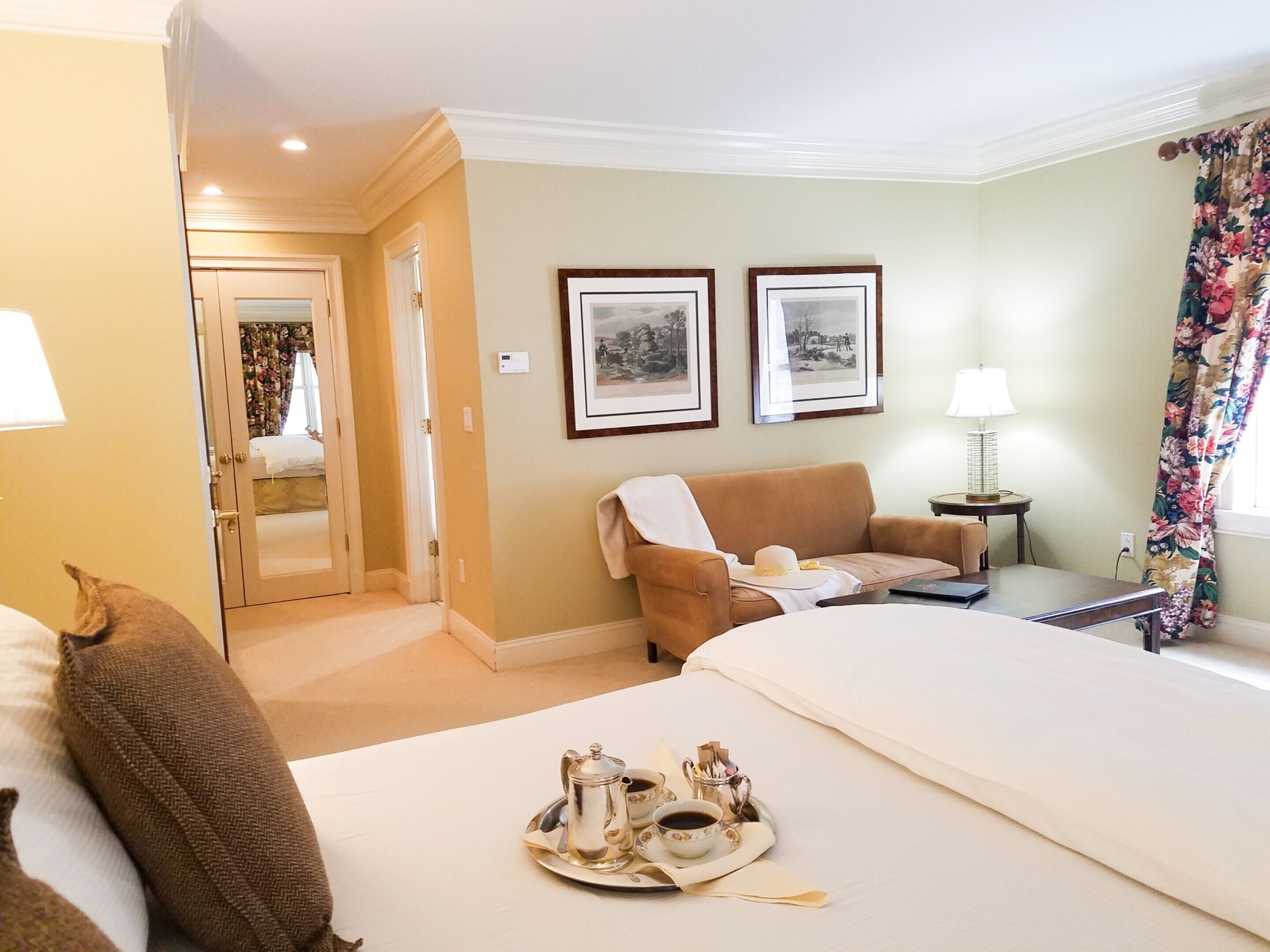 A hotel room with a bed, a sofa, a table with a lamp, and two framed pictures on the wall.