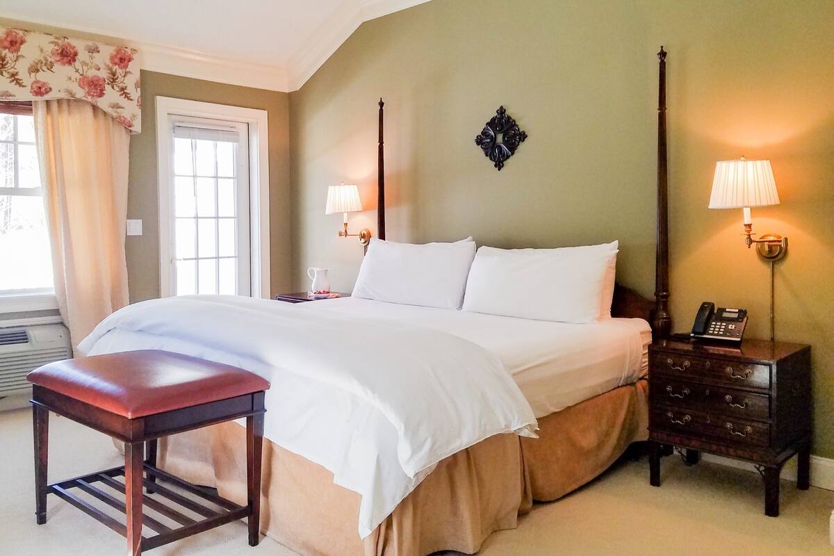 A neatly made bed with white linens, a wooden nightstand with a lamp, and a small table with a chair.