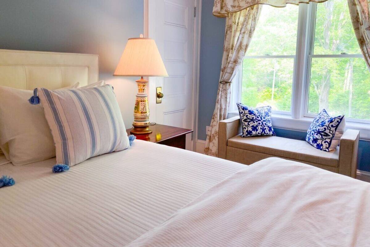 A bedroom with a bed, a nightstand, a lamp, a window with curtains, and a view of greenery outside.