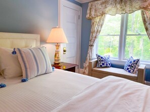 A bedroom with a bed, a nightstand, a lamp, a window with curtains, and a view of greenery outside. - Copper Beech Inn (Ivoryton)