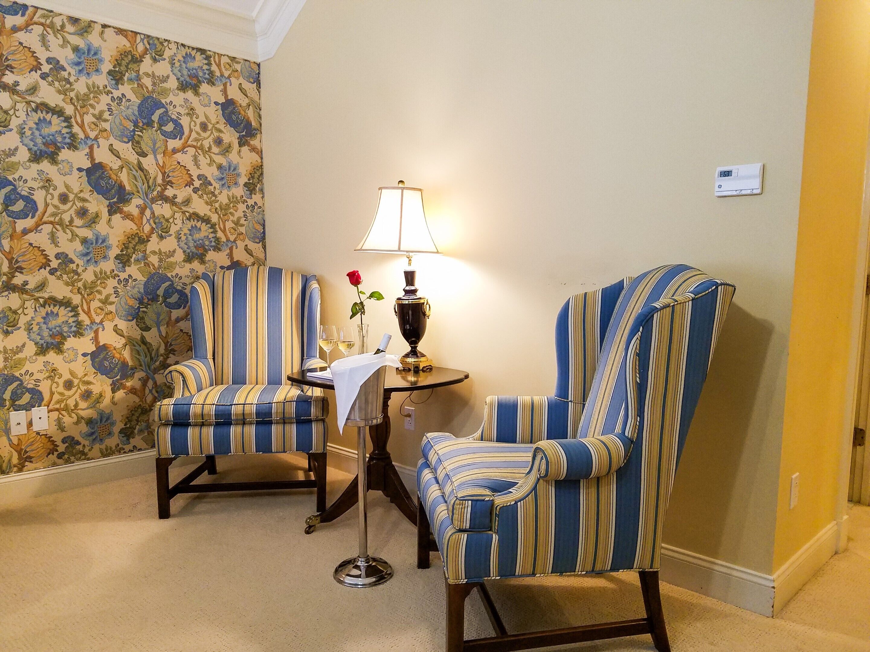 Una habitación con dos sillones a rayas, una pequeña mesa con una lámpara y un papel tapiz floral.