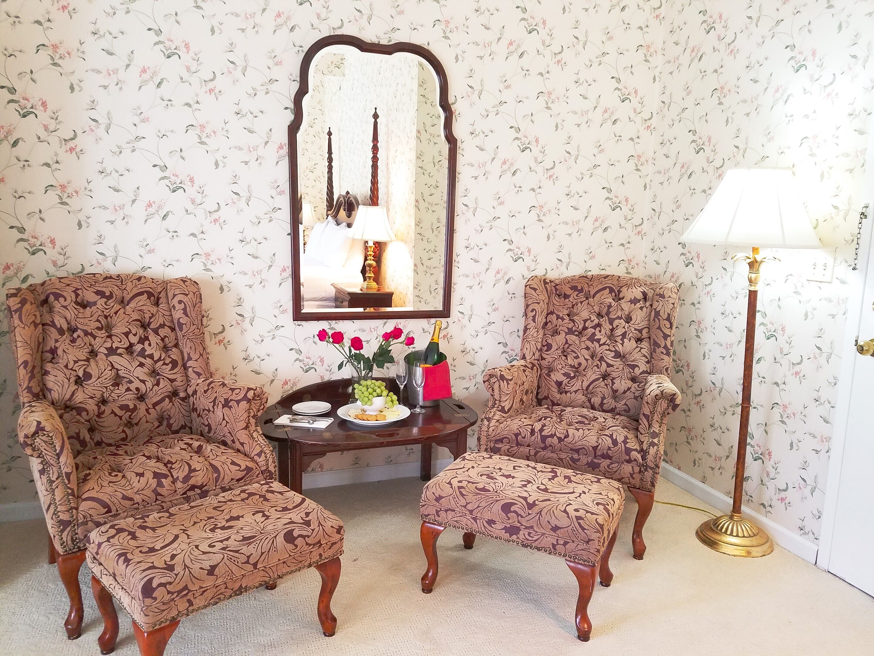 A room with two upholstered armchairs, a small table with a vase of red roses, a lamp, and a mirror on the wall.