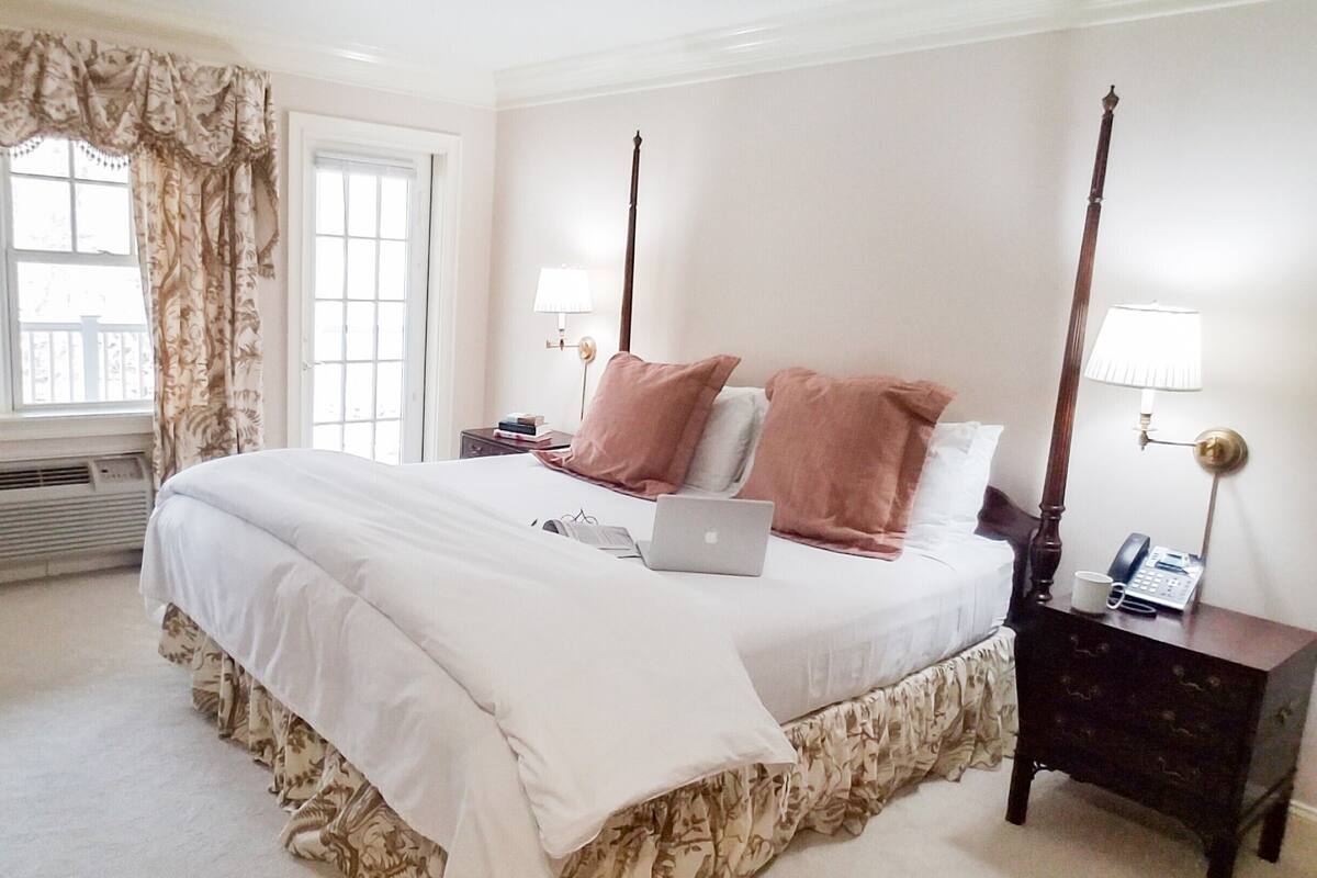 A neatly arranged bedroom with a large bed, a bedside table with a lamp, and a window with curtains.