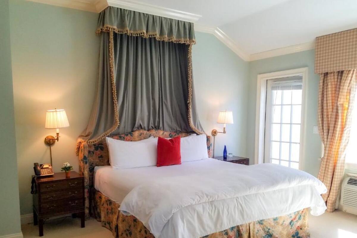 A bedroom with a canopy bed, two bedside tables with lamps, a floral bed skirt, and a window with curtains.