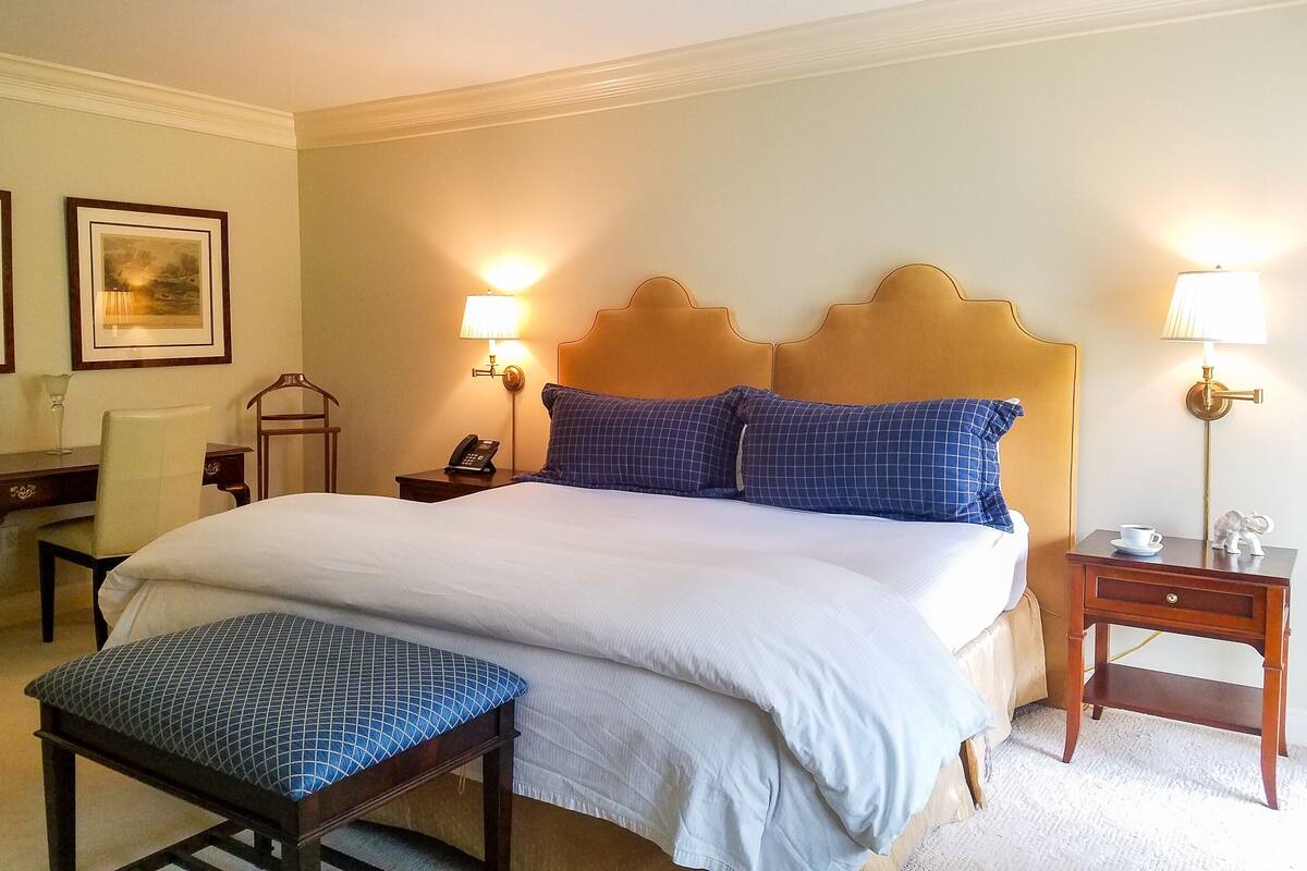 A hotel room with a large bed, two bedside lamps, a bedside table with a phone, a chair, and a small table with a cup.