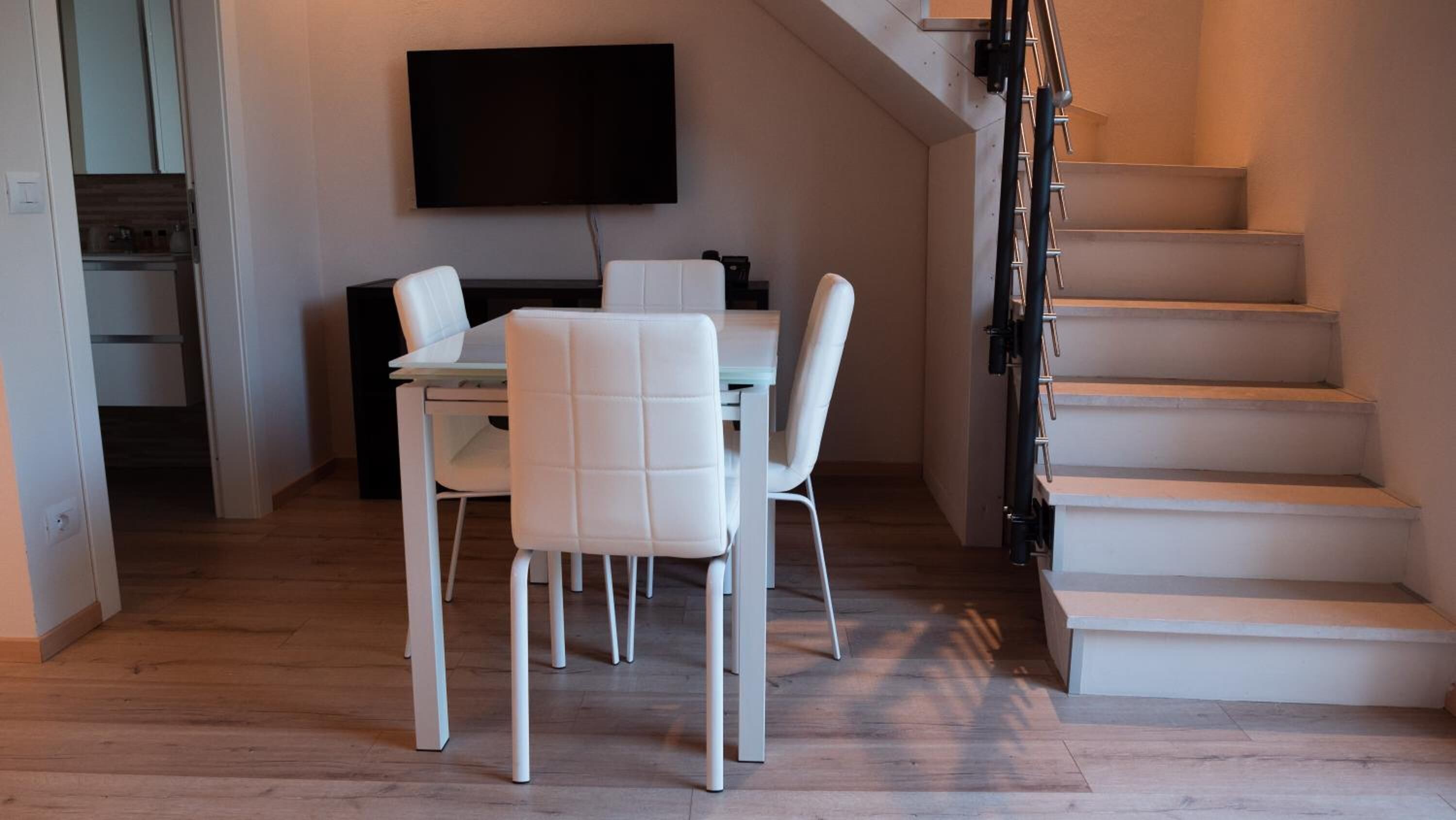 A modern living space with a dining area, a flat-screen TV mounted on the wall, and a staircase leading to an upper level.