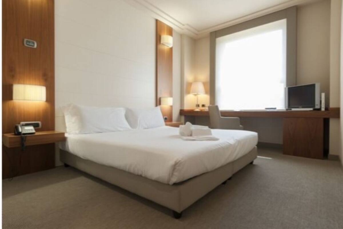 A hotel room with a bed, bedside tables, a desk with a computer, and a window.