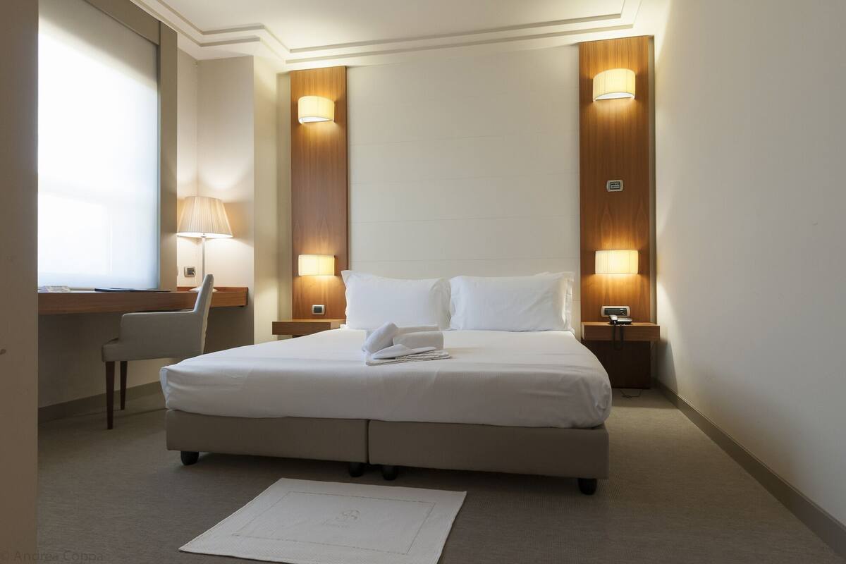 A hotel room with a large bed, two bedside lamps, a desk with a chair, and a window.
