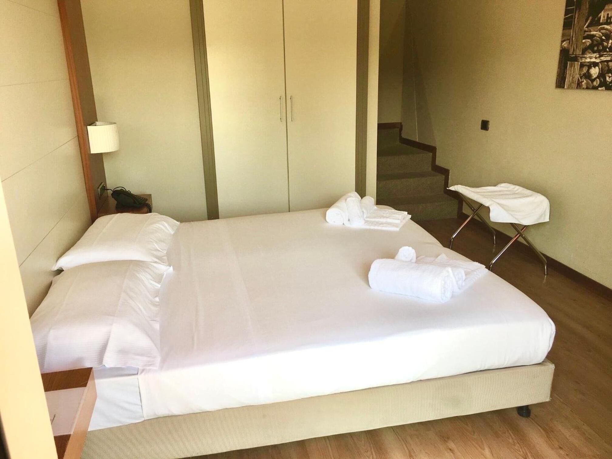 Un letto rifatto con lenzuola e asciugamani bianchi, un comodino con una lampada e una sedia con un asciugamano.