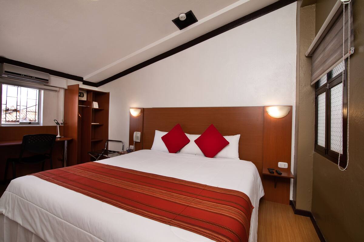 Habitación de hotel con una cama grande, un escritorio y una silla. Se observa una ventana con persianas, un teléfono y una lámpara fijada a la pared.