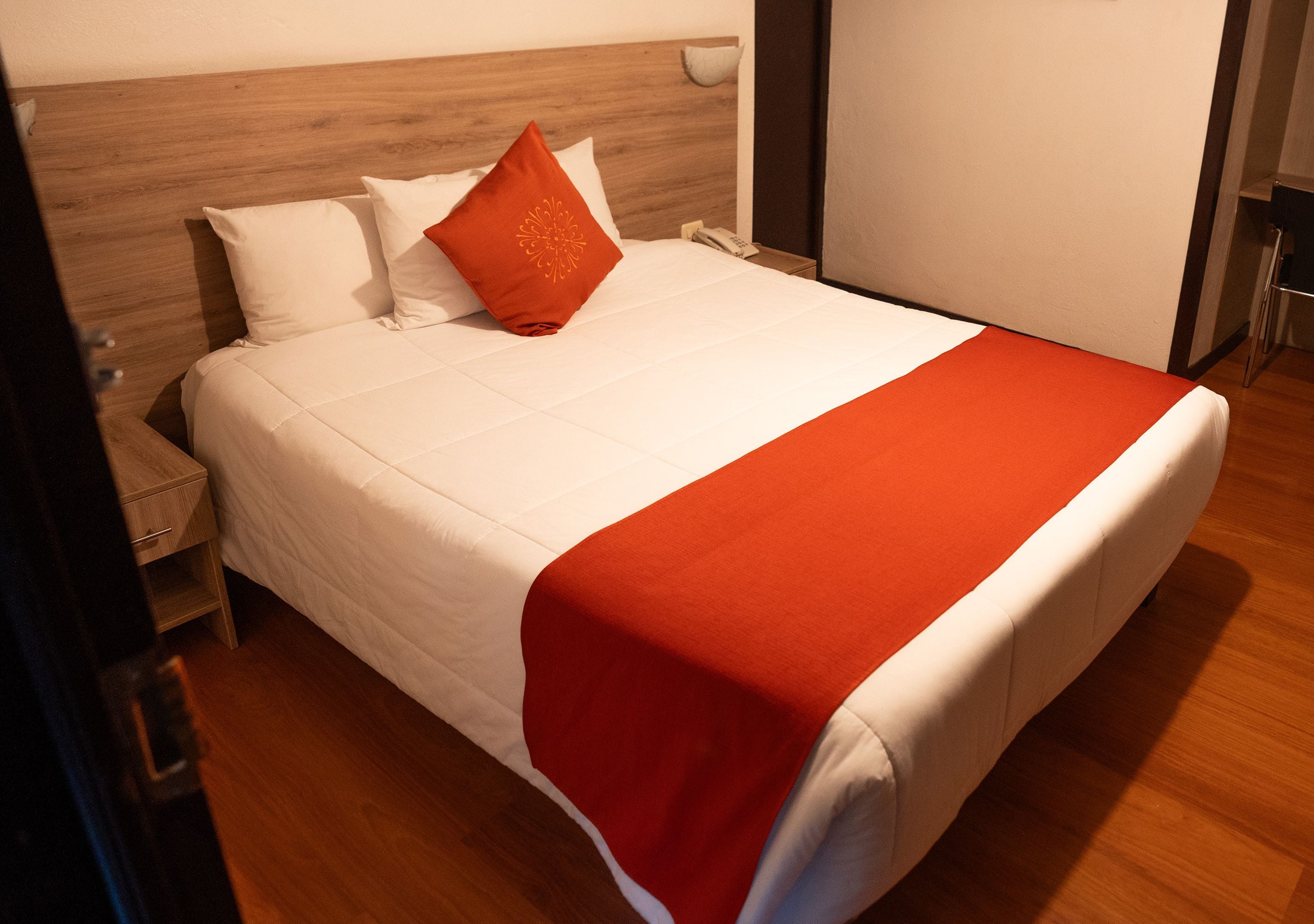 A neatly made bed with white bedding and a red accent pillow, a wooden headboard, and a bedside table with a lamp.