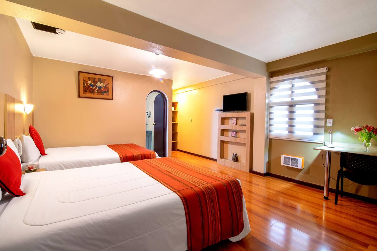 Habitación de hotel con dos camas, un escritorio, un televisor y una ventana con persianas.
