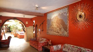 Een kamer met een rode muur, een mozaïekkunstwerk en een zonnespiegel. Er is een bank en een klein tafeltje met een decoratief object erop.