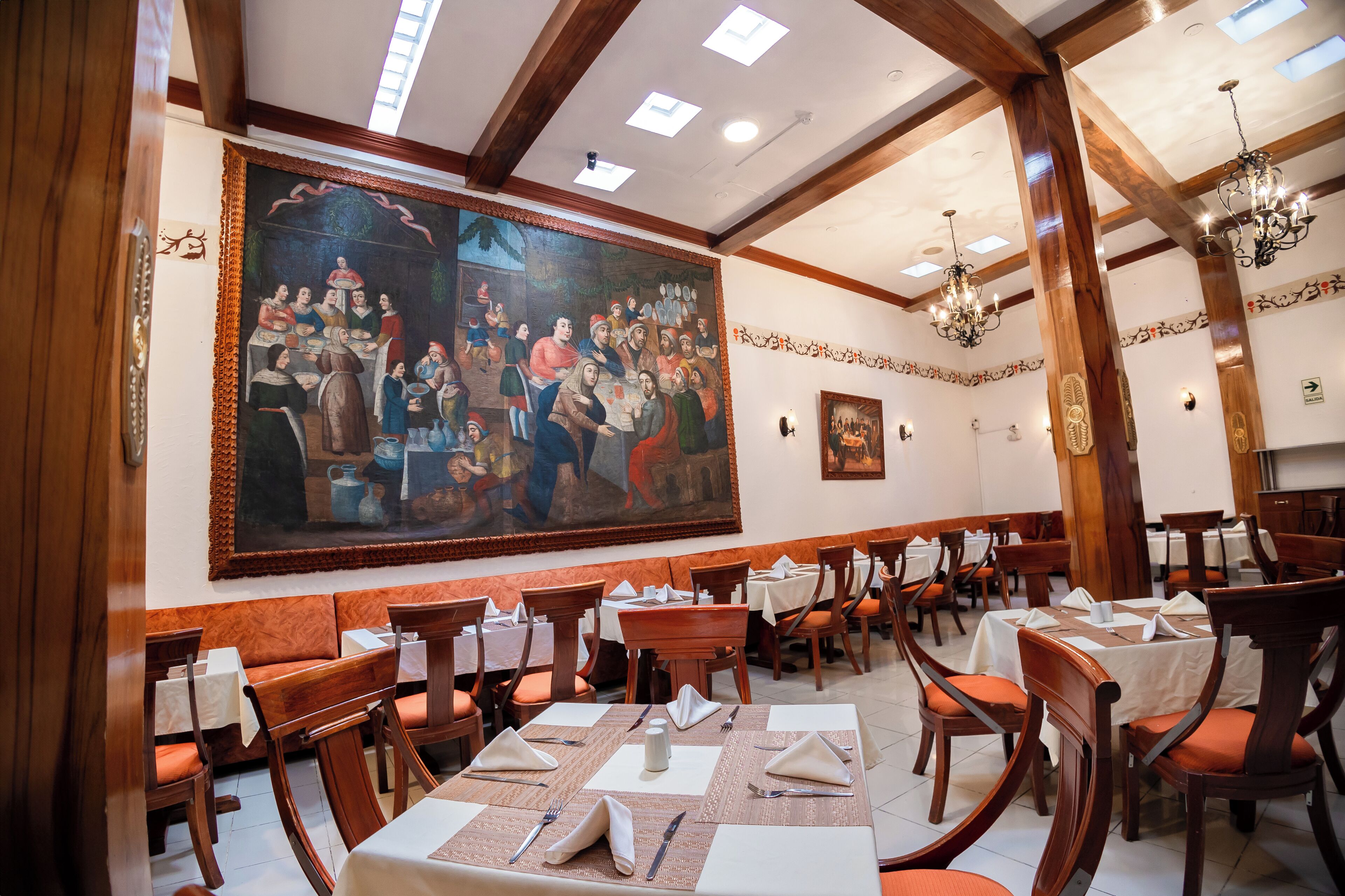 Un espace repas avec des tables et des chaises en bois, une grande fresque murale et un lustre.