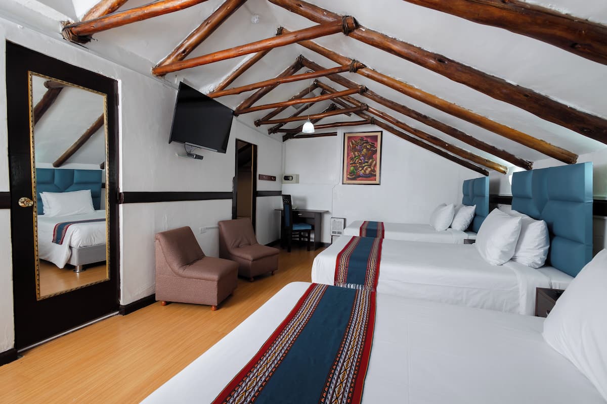 A hotel room with a high ceiling, wooden beams, a bed with white bedding, a blue headboard, a brown armchair, a desk with a chair, and a flat-screen TV mounted on the wall.