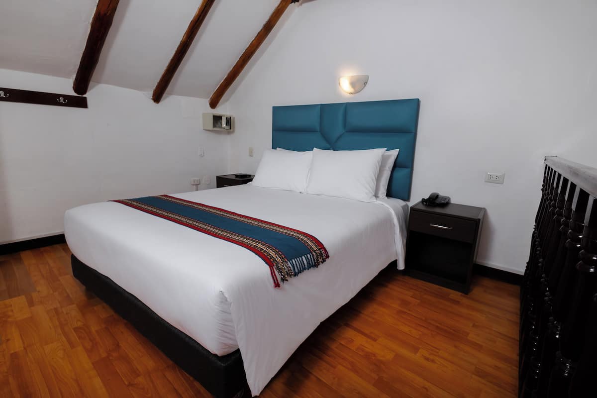 A bedroom with a large bed, wooden flooring, a teal headboard, white bedding, a bedside table with a lamp, and a nightstand with a phone.