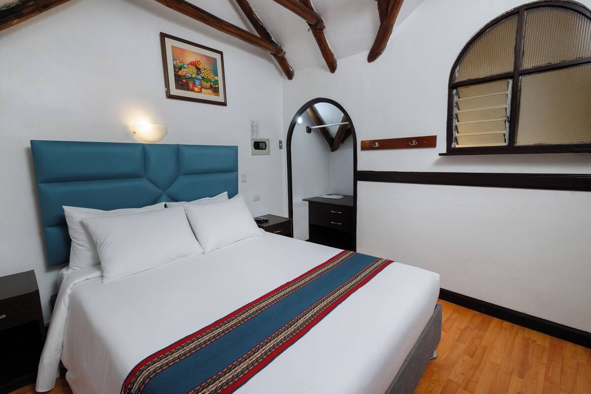 A neatly made bed with a blue headboard, a bedside table, a nightstand, a wall-mounted mirror, and a window with a wooden frame.