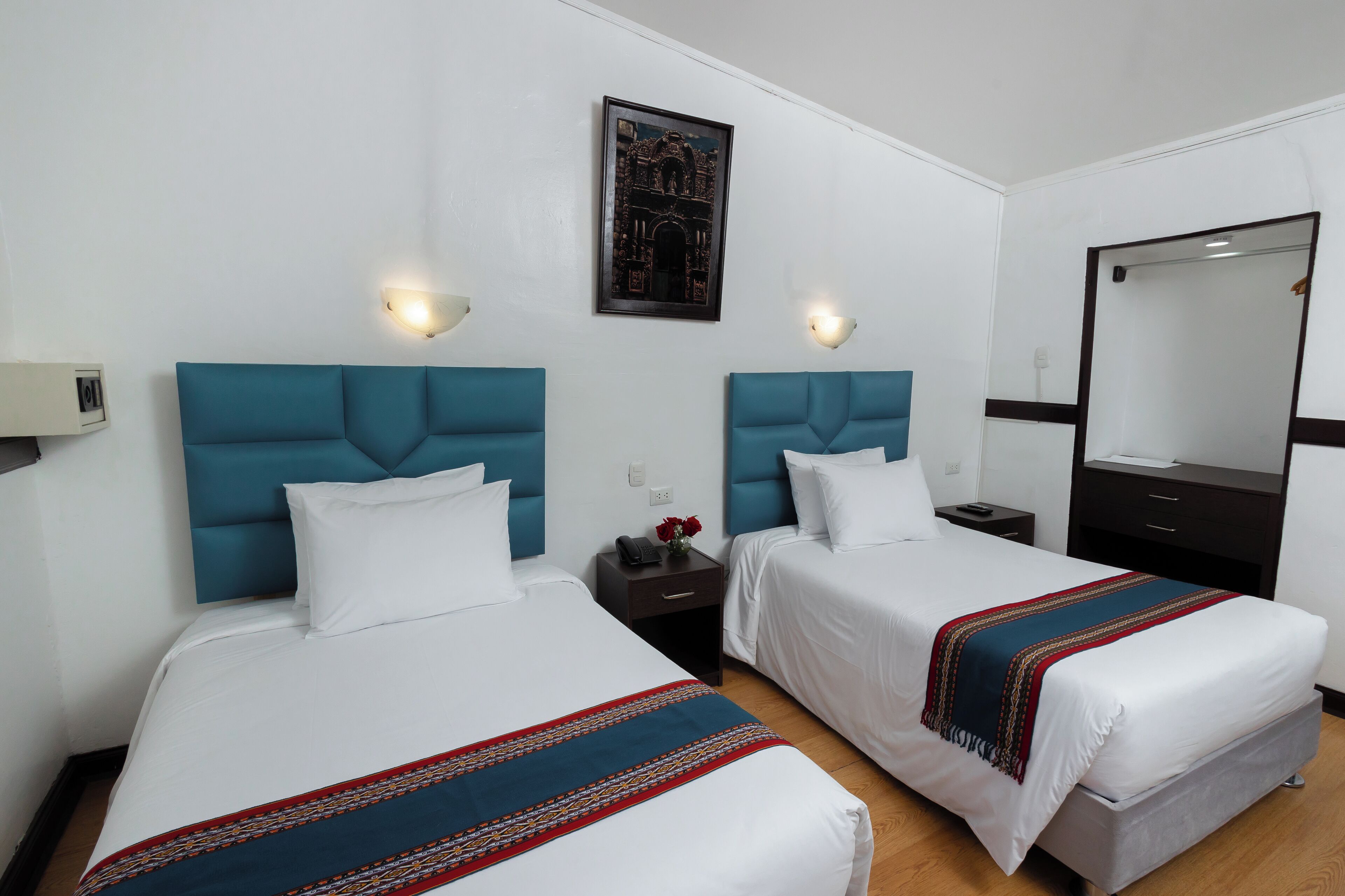A hotel room with two beds, each with a blue headboard, white bedding, and a red and blue striped blanket. There is a nightstand with a phone and a vase with red flowers. A framed picture hangs on the wall above the beds, and a mirror is on the right side of the room.