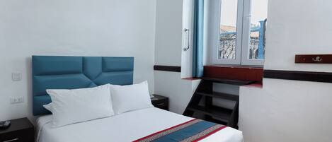A hotel room with a bed, a nightstand, a window with curtains, and a view of the outside.