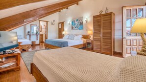 Un dormitorio acogedor con techo inclinado, muebles de madera, cama con manta azul, mesita de noche con lámpara y televisor sobre un mueble de madera.