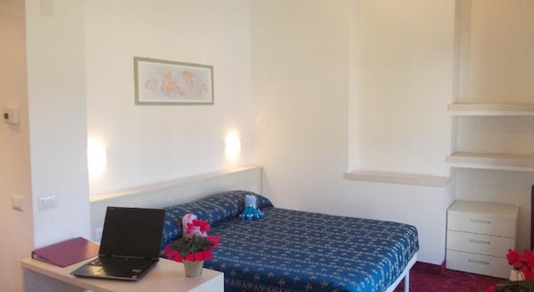 Un dormitorio con una cama, un escritorio con una computadora portátil, una planta pequeña y una mesita de noche.