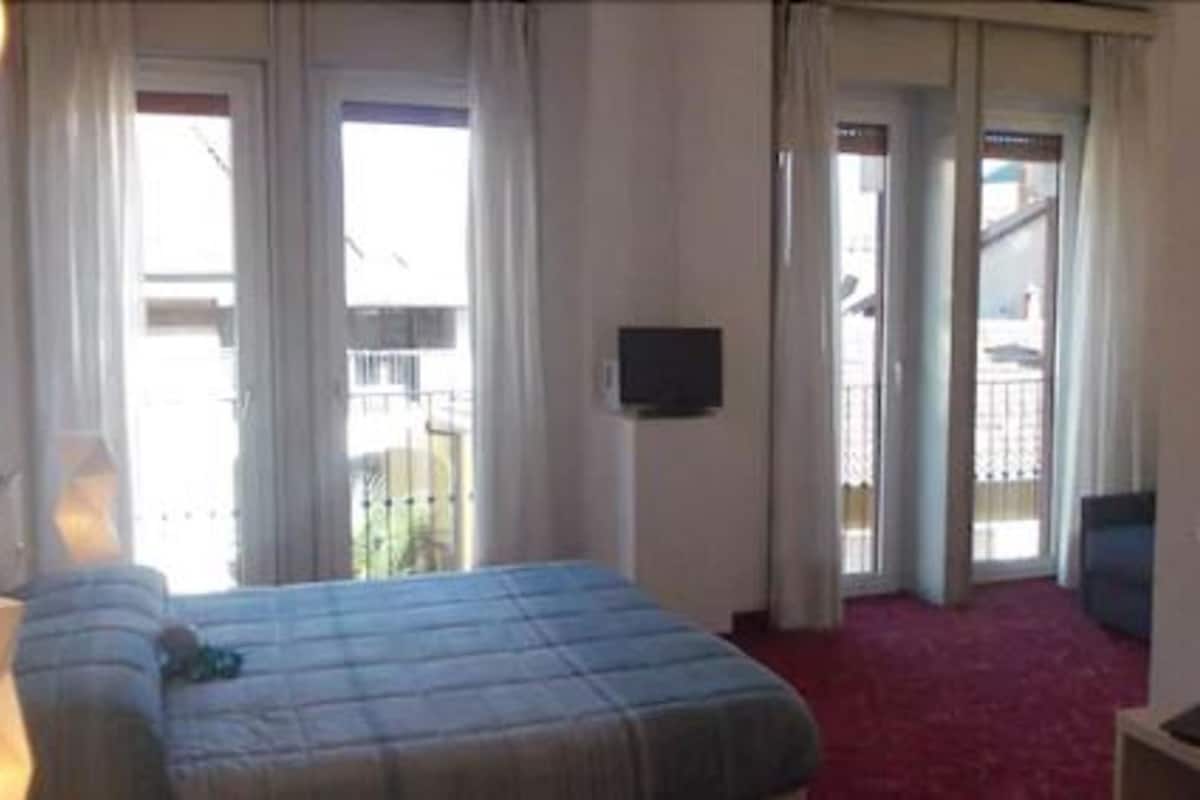 A hotel room with a bed, a TV, and a balcony with curtains.