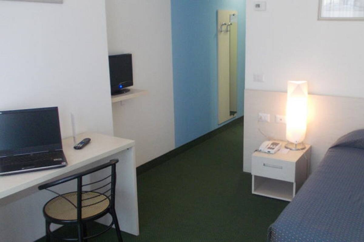 A hotel room with a bed, a desk with a laptop, a chair, a bedside table with a lamp, and a mirror.