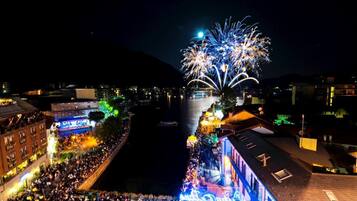 Une vue nocturne d’une ville, avec une foule nombreuse rassemblée le long d’une promenade au bord de l’eau, illuminée par des jeux de lumière colorés et des feux d’artifice.
