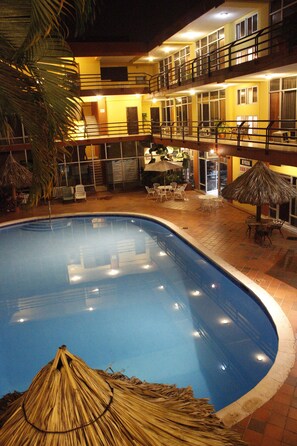 Una piscina con techo de paja, rodeada de mobiliario exterior y un edificio de varios pisos con balcones.