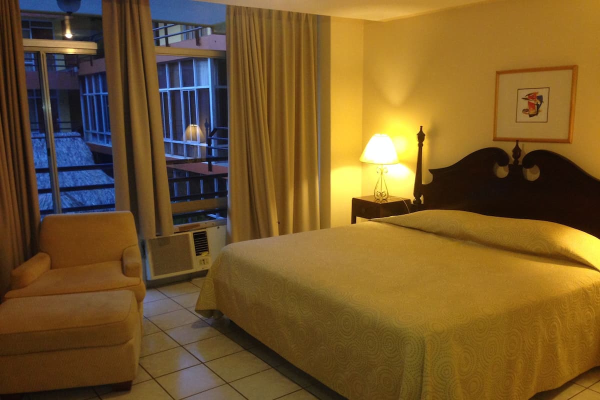A hotel room with a bed, a chair, a window with curtains, a lamp, and a picture on the wall.