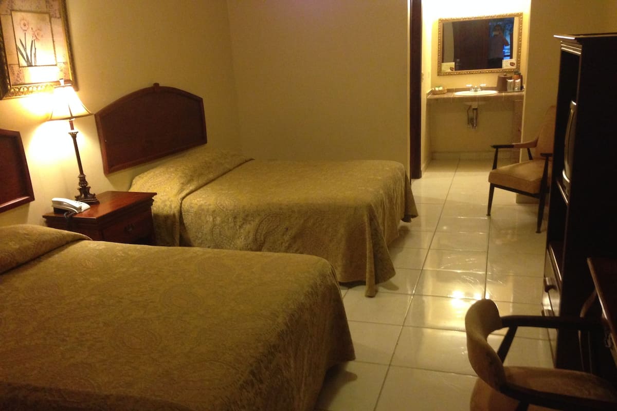 A hotel room with two single beds, a bedside table with a lamp, a chair, and a small desk with a mirror and a sink.