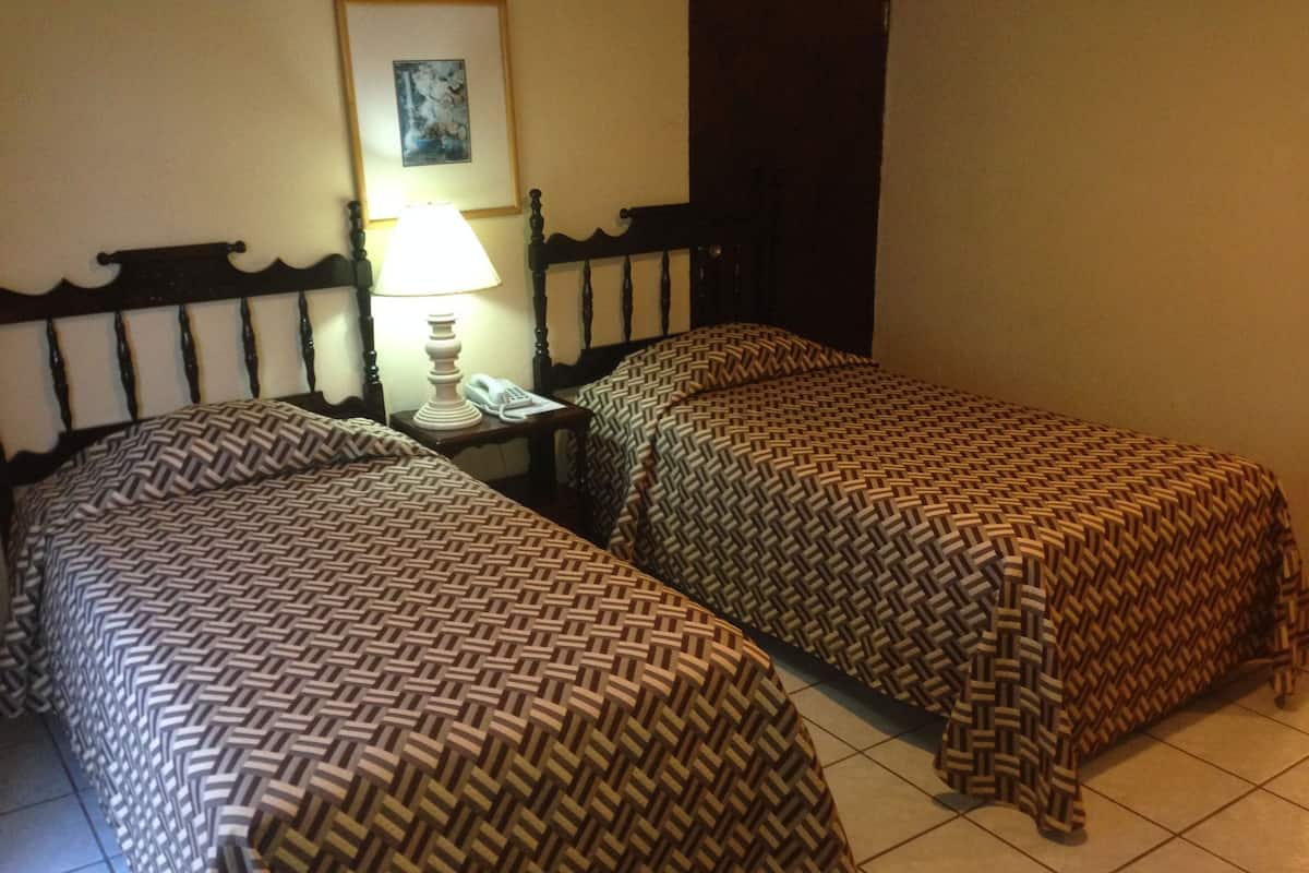 A room with two beds, each with a patterned bedspread, a bedside table with a lamp, and a framed picture on the wall.