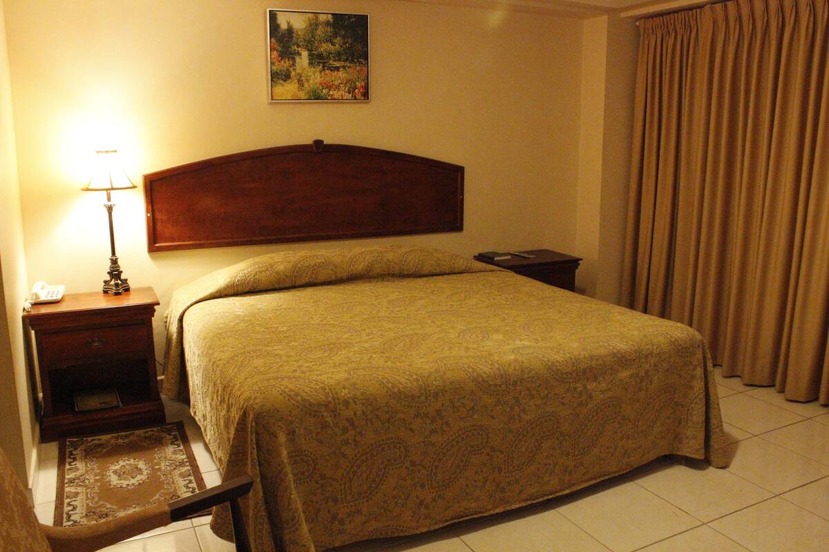 A hotel room with a bed, a nightstand with a lamp, a painting on the wall, and a window with curtains.