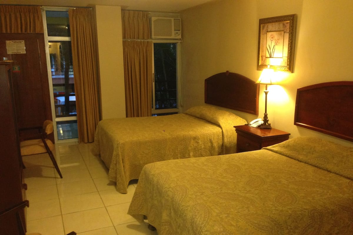 A hotel room with two beds, a chair, a desk with a lamp, and a window with curtains.