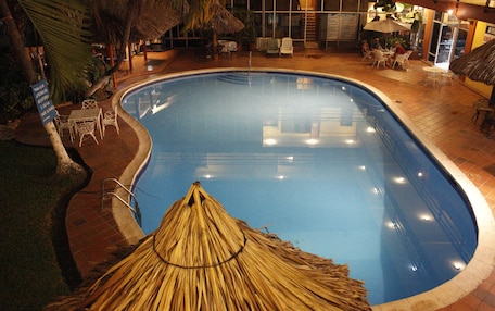 An indoor swimming pool with a thatched roof structure, surrounded by lounge chairs and tables.