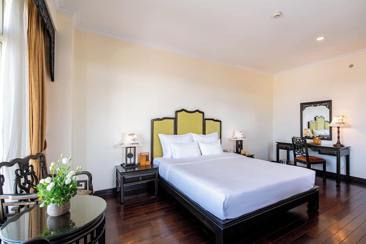 A hotel room with a large bed, a desk, a chair, a table with a vase of flowers, and a mirror on the wall.