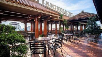 An outdoor café with wooden tables and chairs, surrounded by potted plants and a tiled floor.