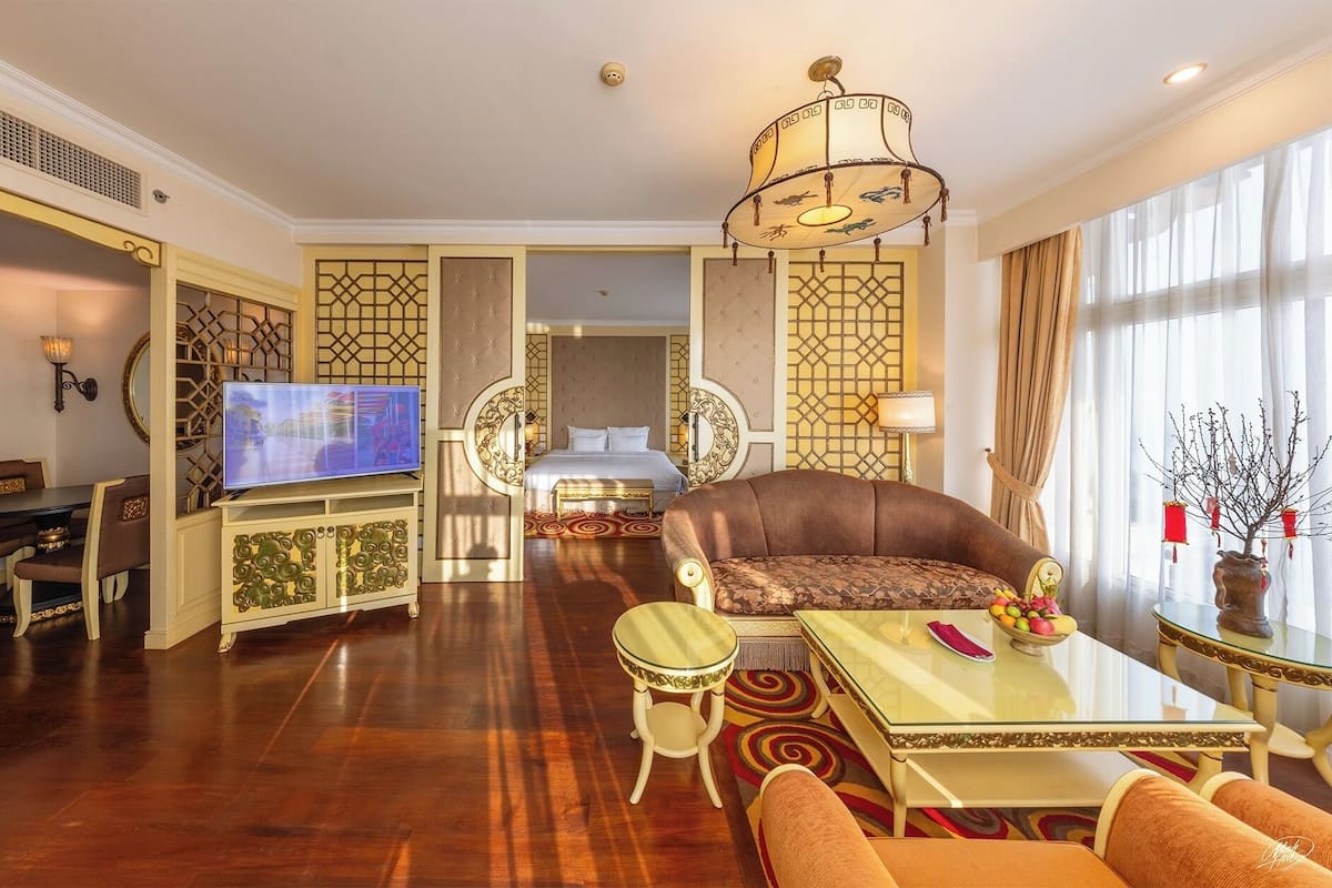 A spacious hotel room with a large television, a comfortable sofa, a dining area, and a decorative chandelier.