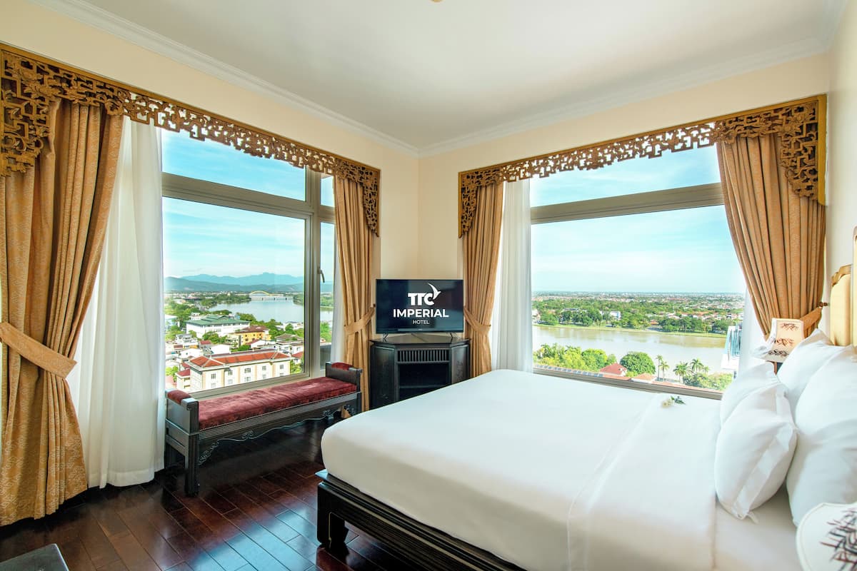 A hotel room with a large bed, a TV on a stand, and a view of a cityscape through large windows.